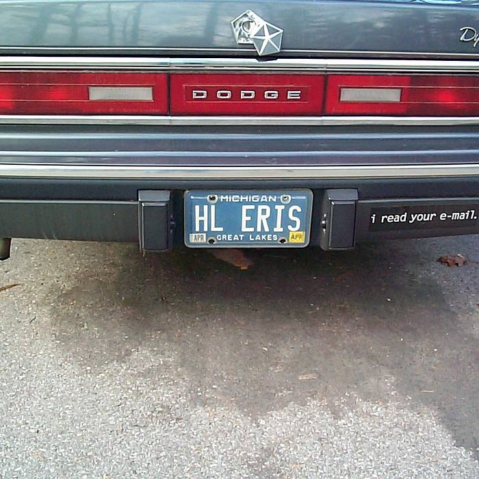 The rear of a dark-colored Dodge Dynasty is shown, featuring a Michigan license plate reading HL ERIS and a black bumper sticker that says 'i read your e-mail.' The car is parked on asphalt with scattered leaves.