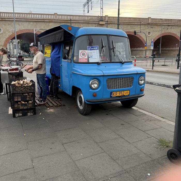 A man grills sausages on a blue vintage food truck parked on a paved sidewalk, with a stone bridge and overhead train lines in the background. Another person stands inside the truck's service window, and a stack of firewood is visible beside the grill.