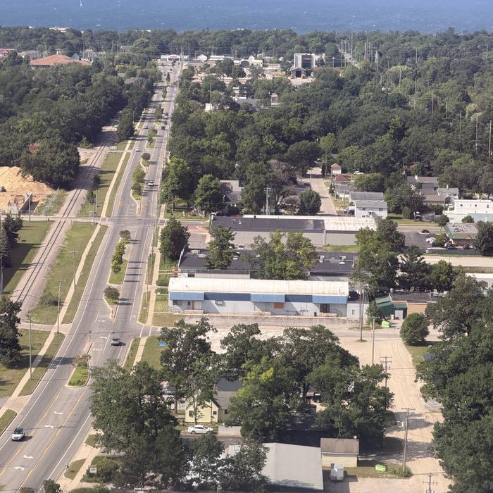 An aerial view of a road running through a suburban area with trees, buildings, and a large body of water in the distance. A construction site with a pile of dirt is visible on the left side of the road.