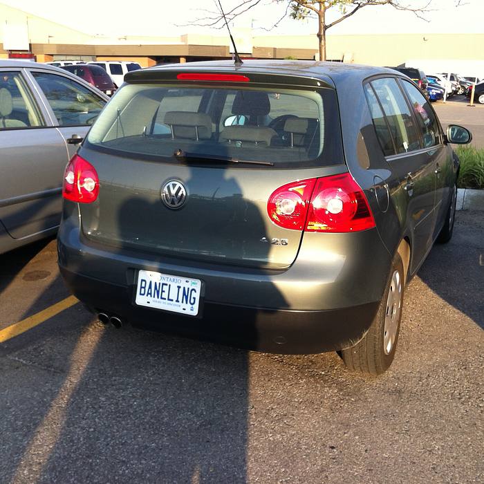 A dark gray Volkswagen hatchback is parked in a lot, with a silver car to its left and other vehicles in the background. The car has a license plate reading BANELING and the number 425 on the rear. The scene is set in a parking lot with yellow lines and a building in the distance.